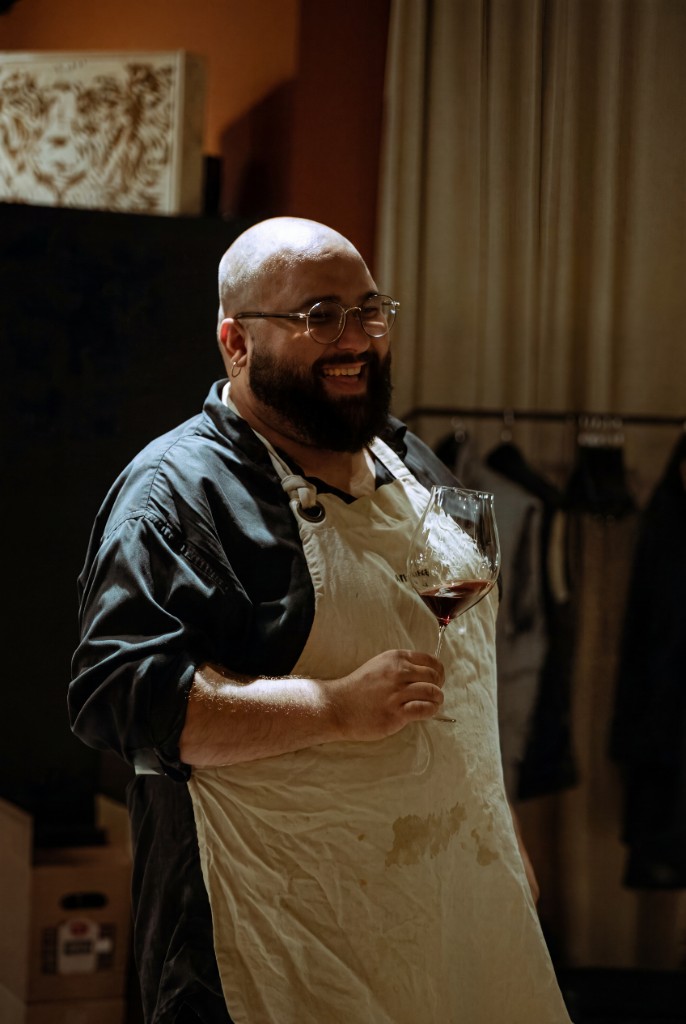 Daniel Gol in a work apron, glass of red wine in hand, mid-laugh behind the counter