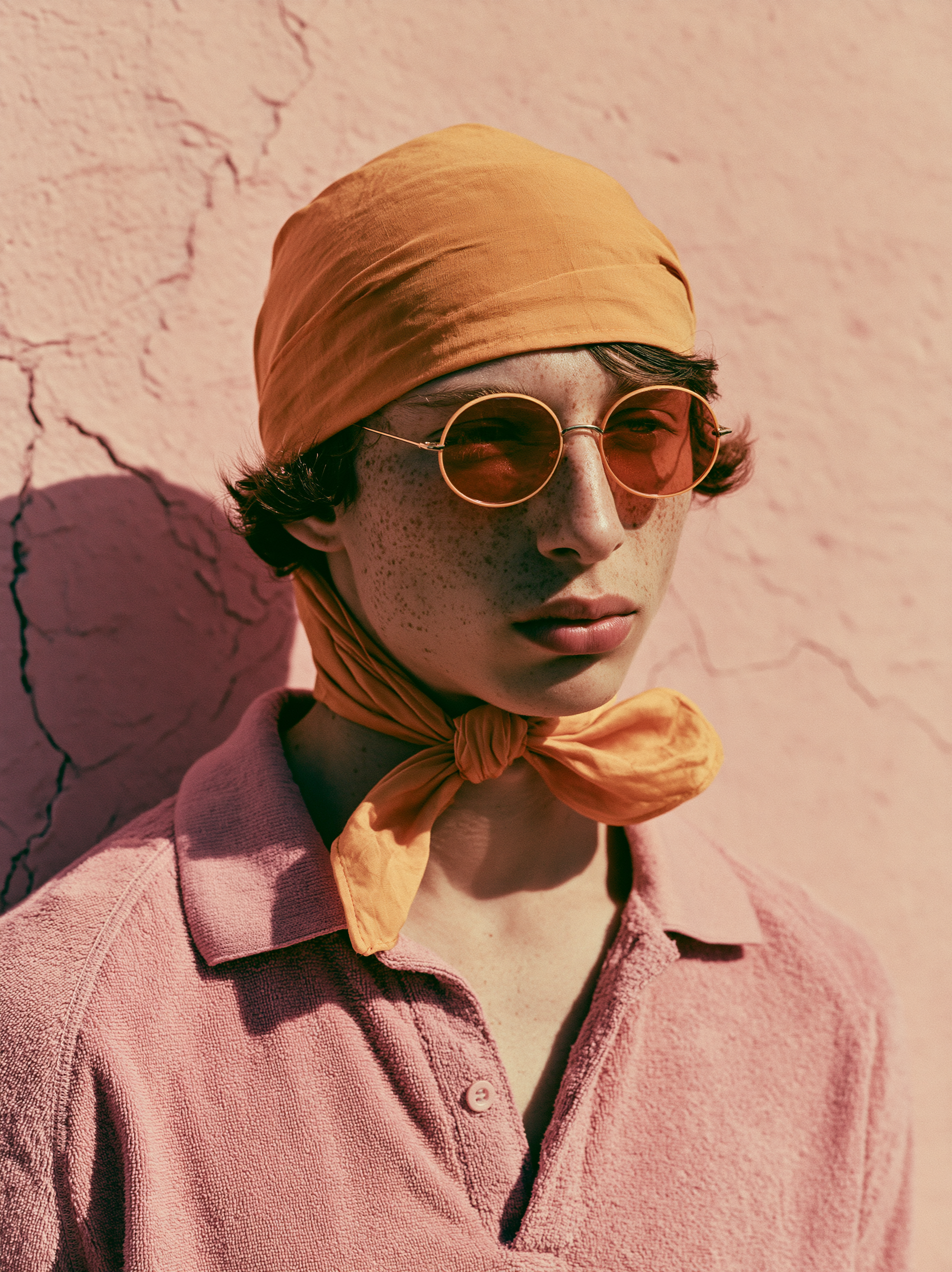 Boy in an orange headscarf and round tinted glasses against a cracked pink wall