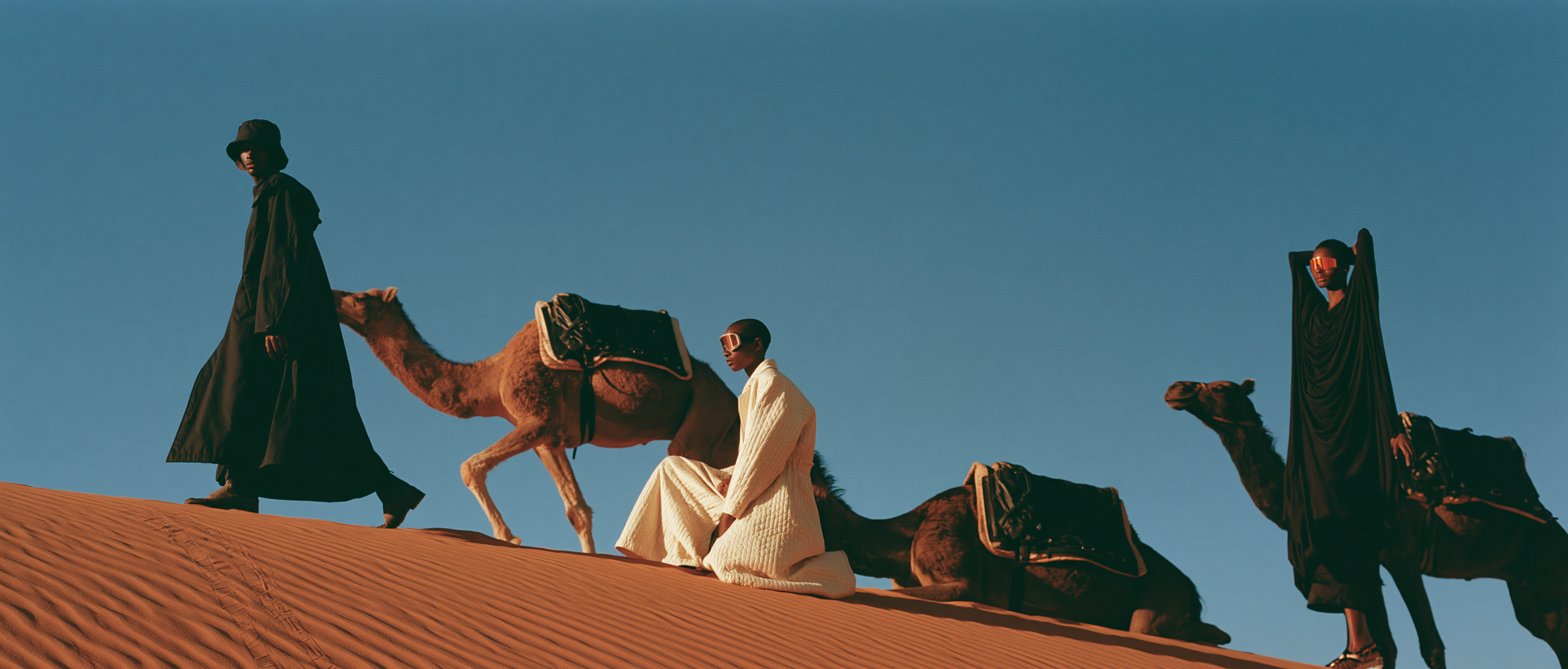 Three models and camels crossing the ridge of a dune