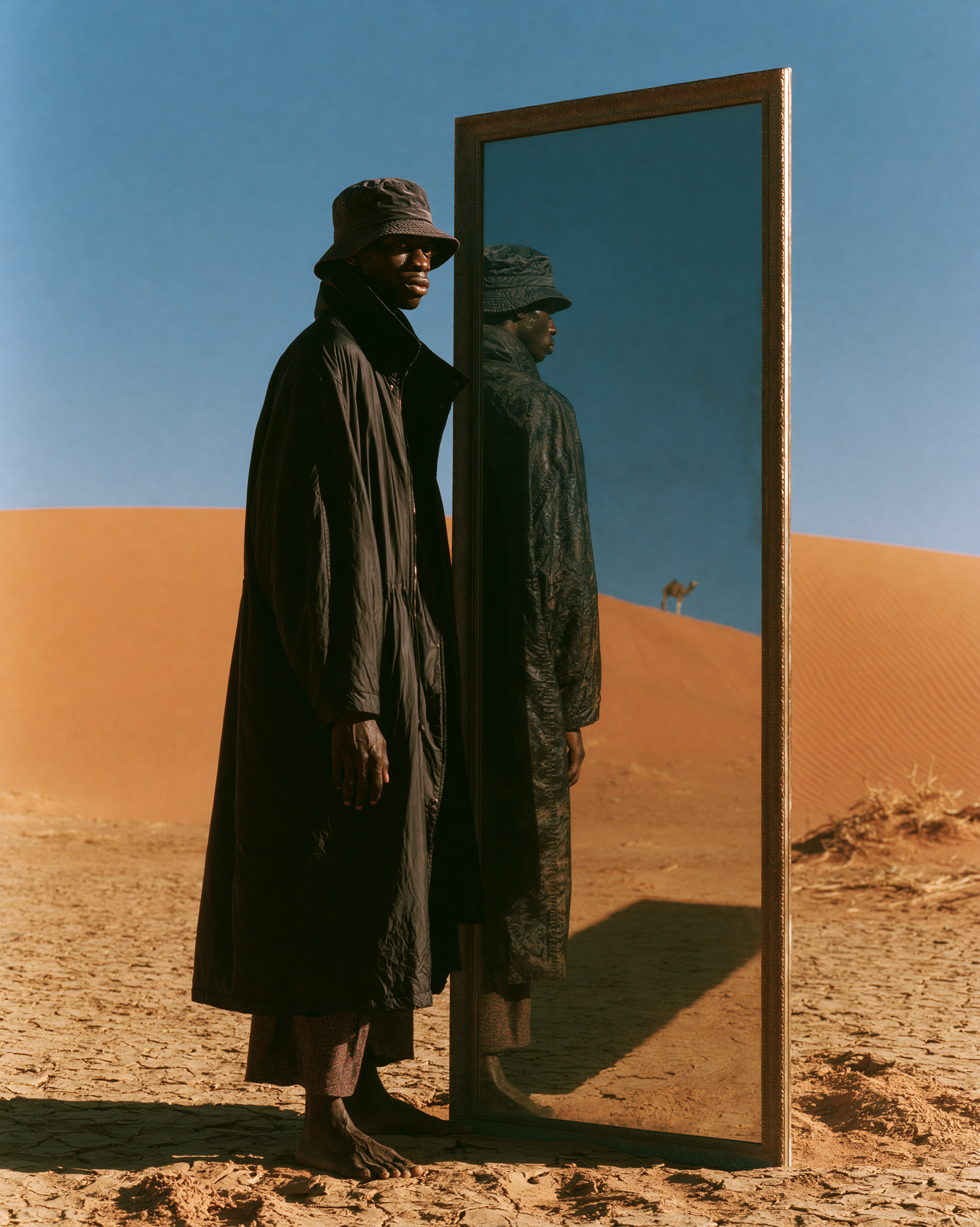 A model beside a free-standing mirror in the desert, a camel on the dune in the reflection