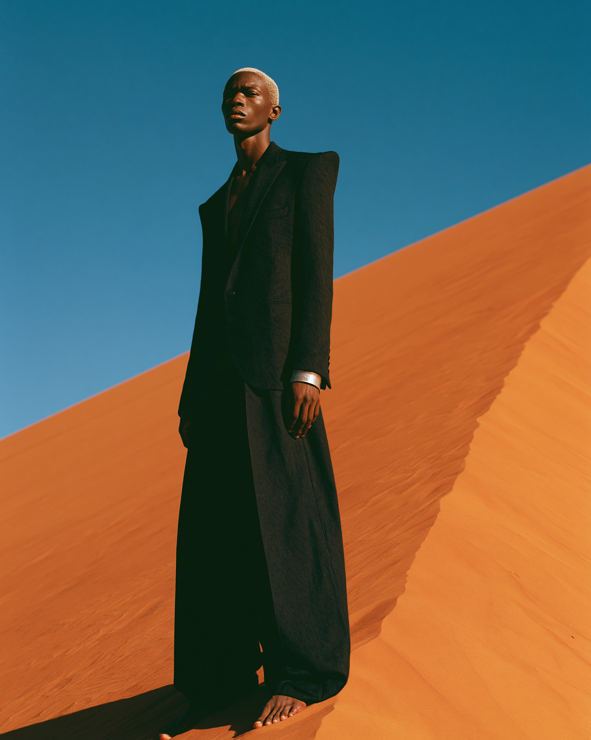 A model in a sharp black suit standing on the crest of a dune against a blue sky
