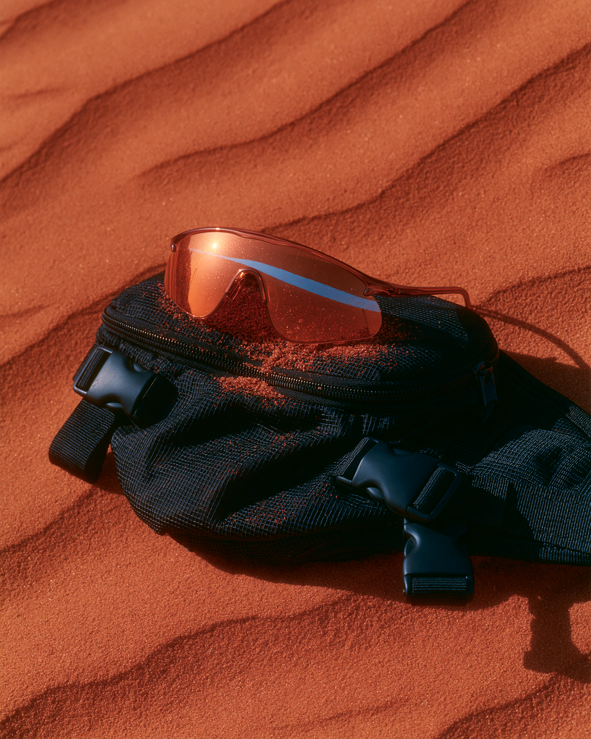 Orange-mirrored sunglasses resting on a black mesh bag in red sand