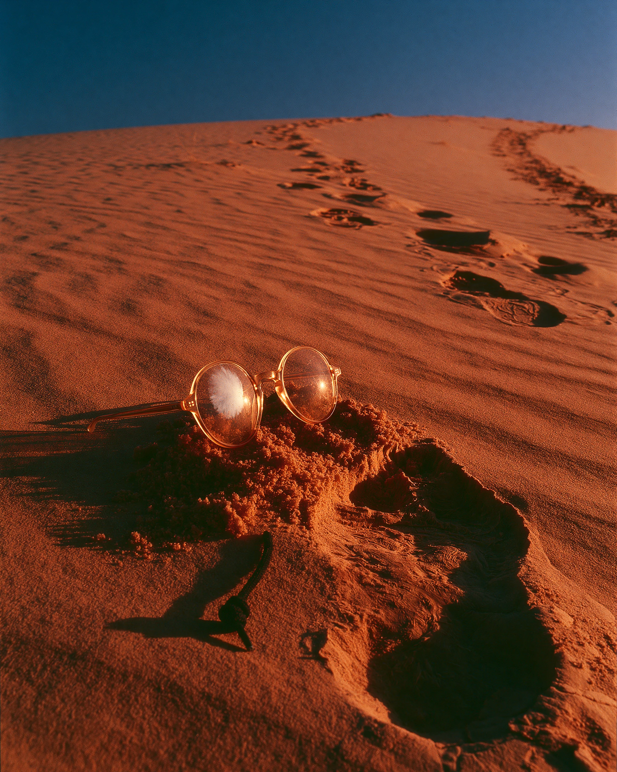 Abandoned amber glasses and a trail of footprints up a dune at sunset