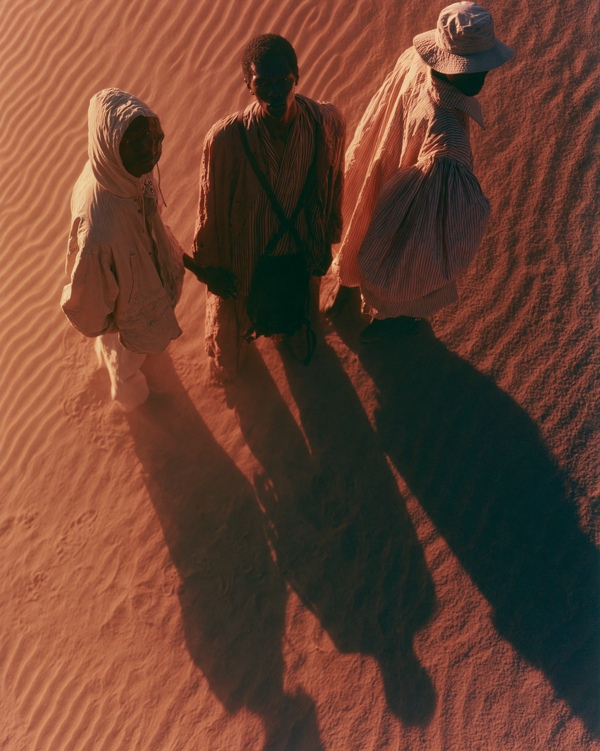 Three figures holding hands seen directly from above, shadows raking the sand