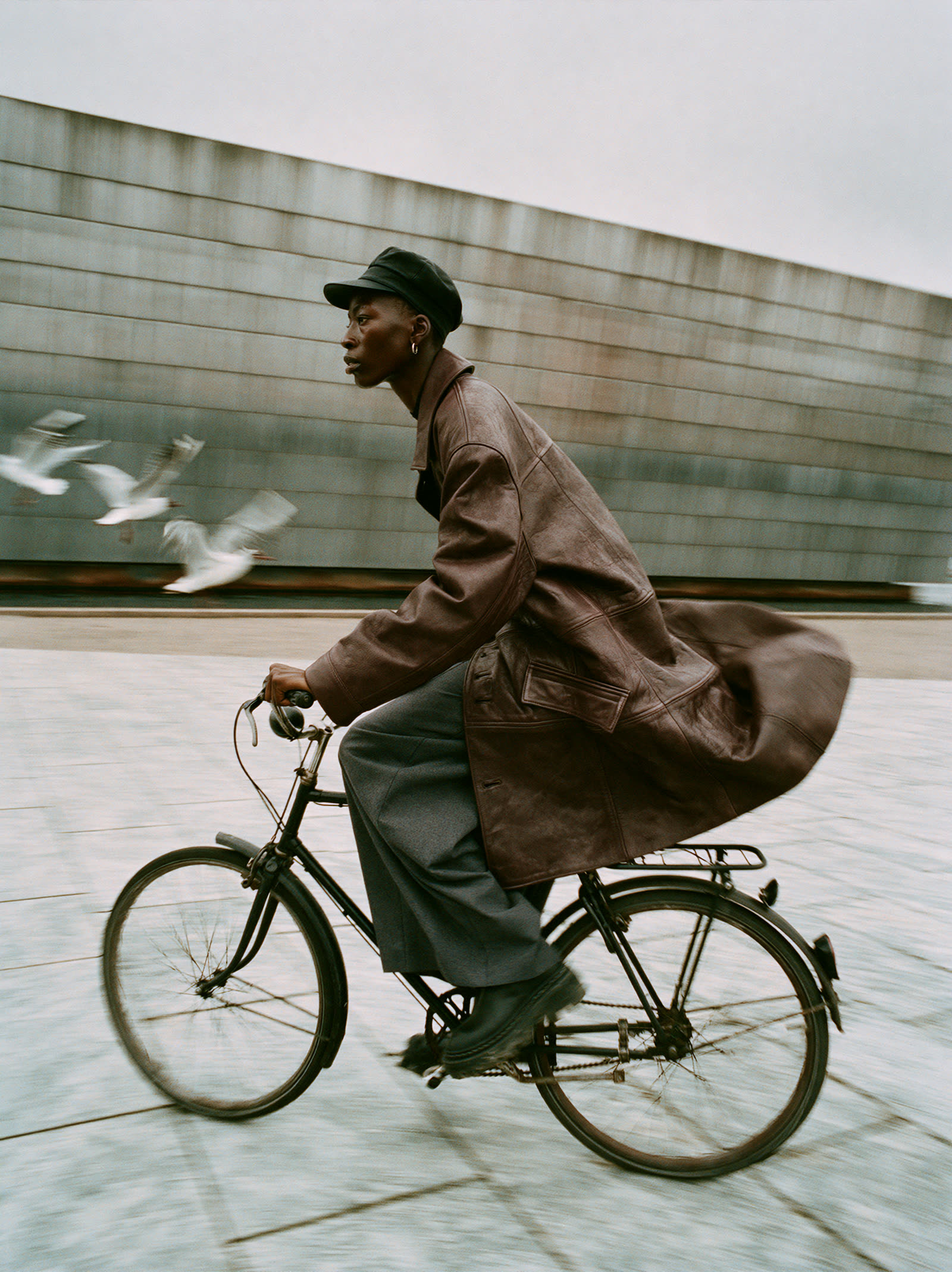 A man in a leather coat cycling past a concrete wall